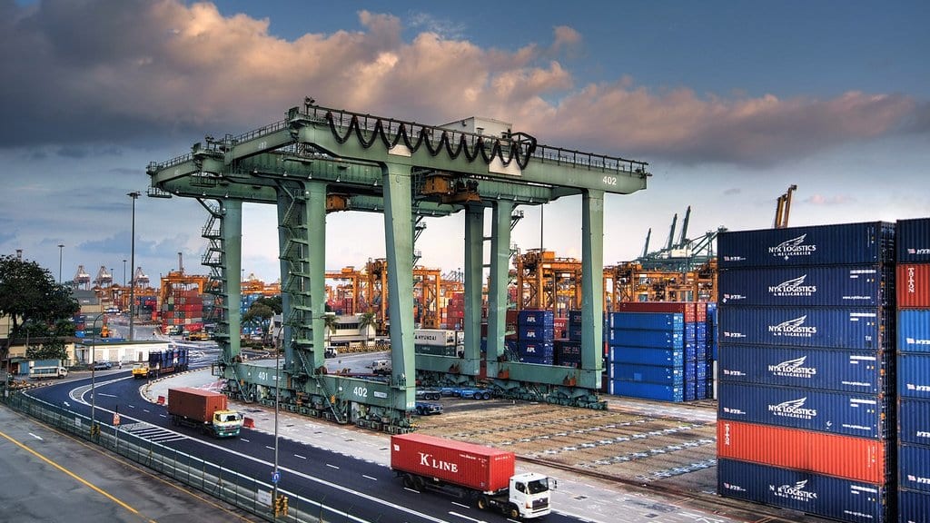A tall gantry crane straddles rows of freight containers at a port container terminal at dusk, with trucks on the service road alongside and more cranes in the distance.