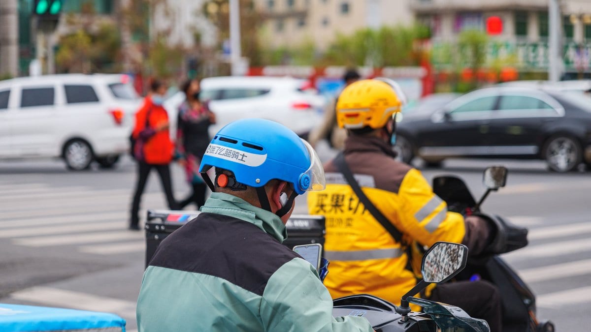 Two food delivery couriers on electric scooters wait at a city intersection, insulated delivery boxes mounted behind their seats.