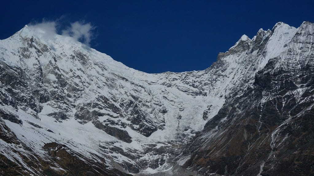 A steep Himalayan face of bare rock and hanging ice under a deep blue sky, with gray glacier debris spread across its base.