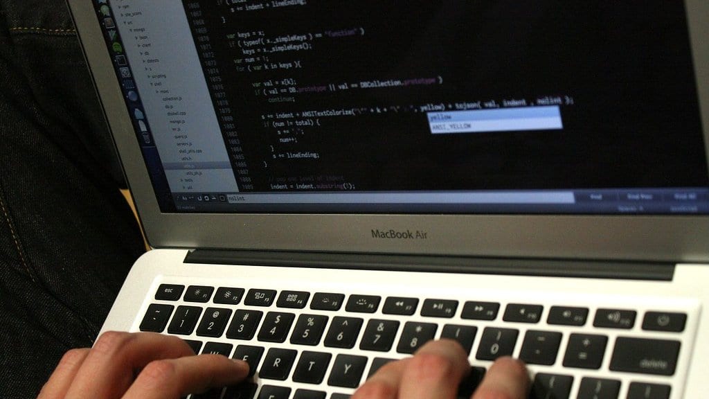 Close-up of a person's hands typing on a laptop keyboard, with lines of program code displayed on the screen above.