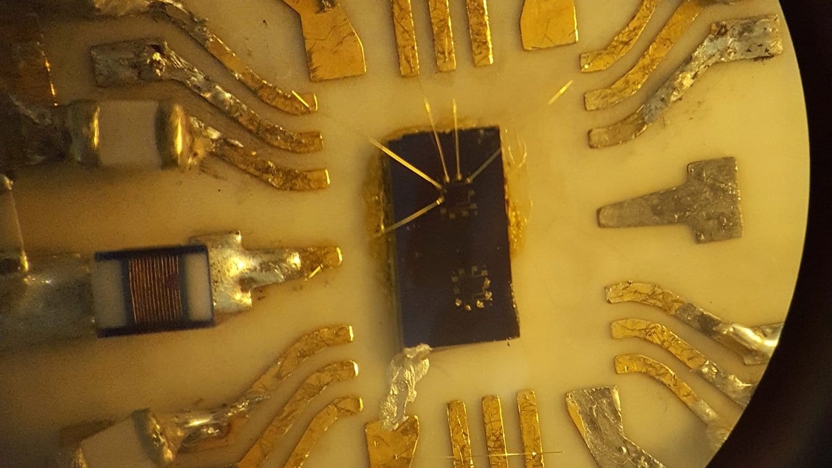 A small dark semiconductor chip at the center of a gold chip carrier, linked by fine bond wires to gold contact pads, photographed through a microscope.