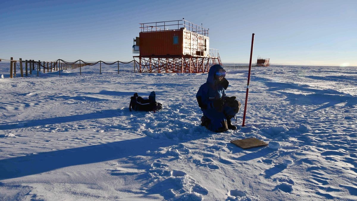 A researcher in a blue parka kneels on wind-rippled snow beside a marked measuring pole, with an Antarctic research station raised on stilts on the flat horizon behind.