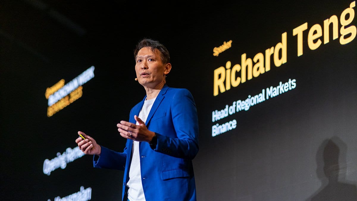 A man in a blue jacket speaks on a dark conference stage; the screen behind him reads Richard Teng, Head of Regional Markets, Binance.